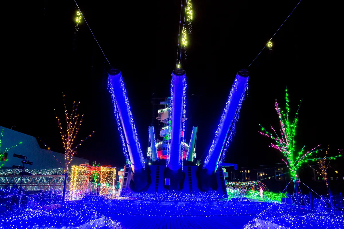 The guns of a Battleship Wisconsin turret lit in blue, with colored lights on trees and a lit gift box around it at night